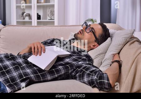 Bookish tranquility: Young man with facial hair and glasses dozes off while reading on couch in living room. Embracing relaxation and literature. Stock Photo