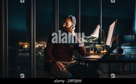 A dedicated businessman works late into the night at his desk, communicating with colleagues using his laptop and phone. The bustling office environme Stock Photo