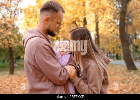 Young mother and father are walking with a newborn baby in the autumn park. Mother kissing newborn daughter Stock Photo