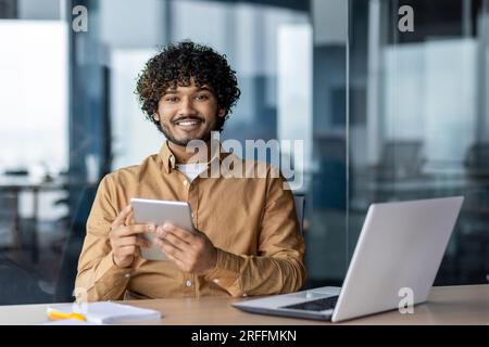 Portrait of young Indian programmer, man working inside company office, using laptop to write software code, businessman smiling and looking at camera, holding tablet computer. Stock Photo