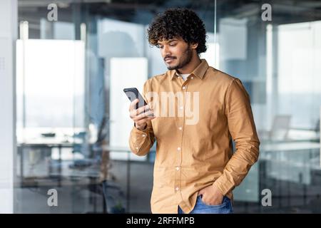 Serious and confident businessman inside office standing near window, hispanic man holding phone in hands thinking reading online message, man using app on smartphone at workplace. Stock Photo