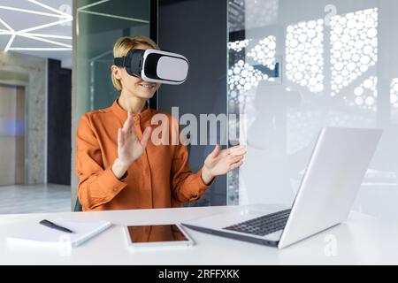 Young beautiful business woman using virtual reality glasses at workplace, employee on online meeting remotely sitting inside office. Stock Photo
