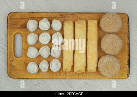 Assorted homemade semi-finished products on wooden board. Frozen cutlets, dumplings and pancake rolls. Preparations of homemade food. Stock Photo