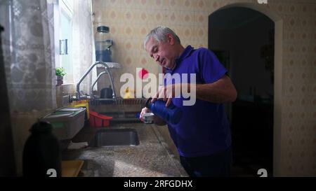 Senior man standing by kitchen sink serving himself coffee and drinking in contemplation staring out window view Stock Photo