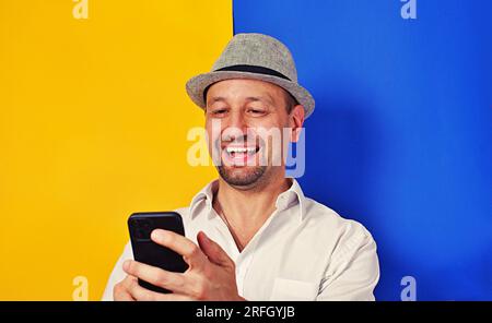 Photo portrait of happy man holding phone in two hands isolated on yellow and blue , two colored background, smile broadly, feel happy Stock Photo