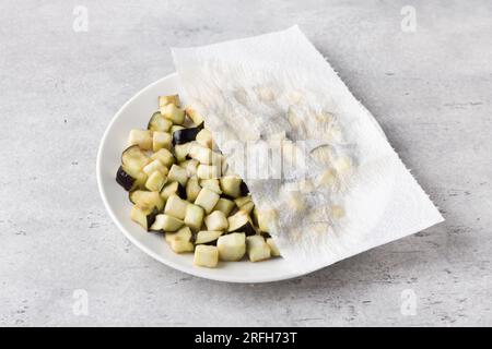 Drying of diced eggplants with a paper towel on a gray textured background. Processing eggplant before cooking, removing bitterness. Stock Photo