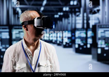Aged computer scientist immersed in virtual reality at data center, doing hardware maintenance. Worker using VR headset to optimize server rigs system performance, harnessing cutting edge technology Stock Photo