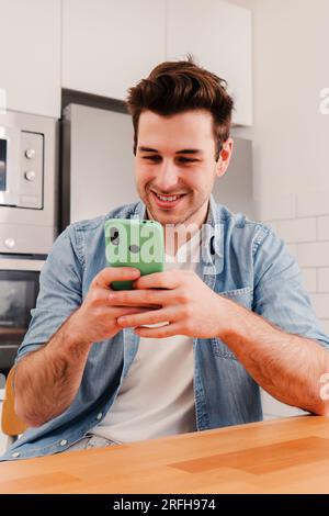 Vertical portrait of happy guy using his smart phone chatting sending messages on social media sitting at home kitchen and taking a break. Smiling young man playing a video game on cellphone. High quality photo Stock Photo