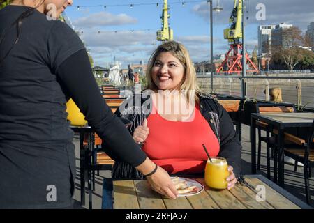 young blonde plus size latin woman smiling very happy, sitting outside the restaurant at sunset, while the waitress serves her a sandwich with orange Stock Photo
