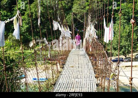 A Bhutanese man wearing the traditional Gho walks across a suspension bridge in rural Bhutan. Faded prayer flags hang from the metal support cables. Stock Photo