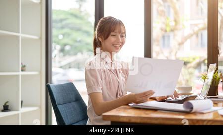A happy Asian businesswoman is examining a financial report, analyzing sales data, and working on paperwork at her desk in the office. Stock Photo