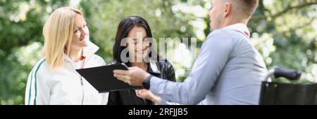 Two female students and one male student study together in park, man is sitting in wheelchair. Stock Photo