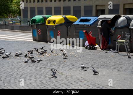 Homeless woman with a cane looking for food in a garbage container on a Barcelona street with her shopping cart and bags next to her, and many pigeons Stock Photo