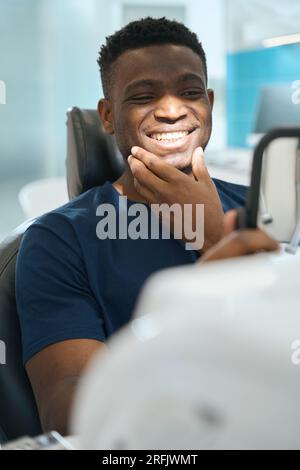 Happy African American man looking at his healthy teeth and smiling, satisfied with qualified stomatological team work, private clinic service Stock Photo