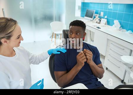 Smiling woman dentist calming down scared young African American man who coming to doctor appointment and sitting in dental chair Stock Photo