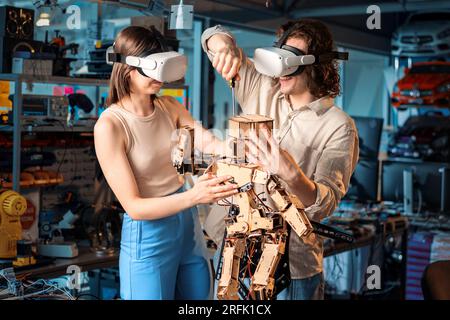 Young man and woman in VR glasses doing experiments in robotics in a laboratory. Wooden robot on the table Stock Photo