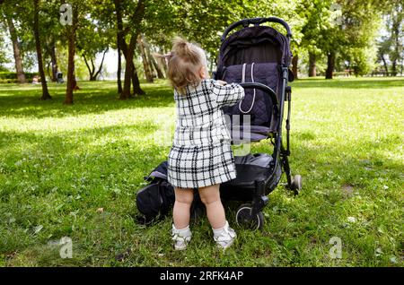 Baby pushing a stroller on a walk in summer park. Adorable little girl near pushchair and waiting for mom Stock Photo