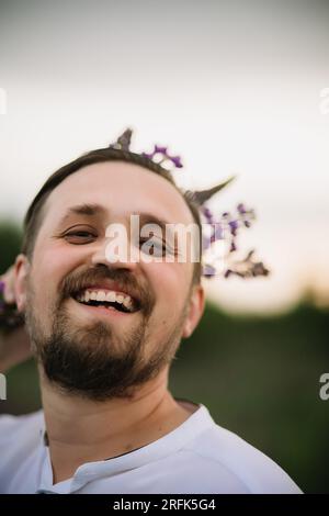 Young smiling man with bouquet of lupines like a crown. Sunset or sunrise, bright evening light. Stock Photo
