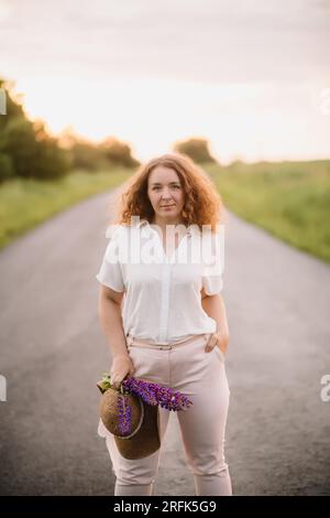 Young woman stands in white shirt on road with purple and pink lupins. Beautiful young woman with curly hair with bouquet of lupins. Sunset or sunrise Stock Photo