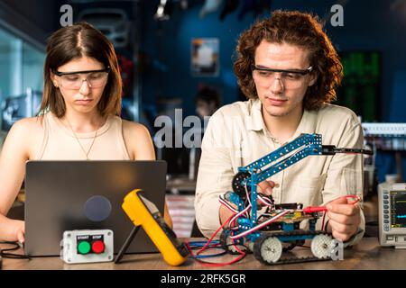 Young man and woman in protective glasses doing experiments in robotics in a laboratory using a computer. Robot and tools on the table Stock Photo