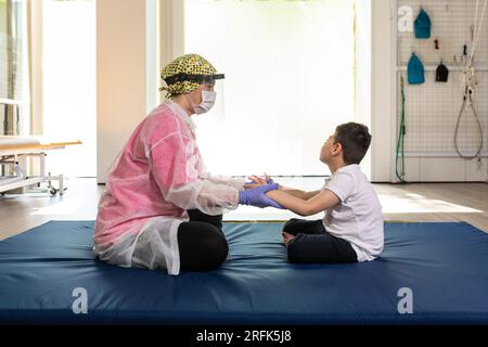 child patient and female physiotherapist in rehabilitation session, exercises on mat during coronavirus, confinement. protective mask. Stock Photo