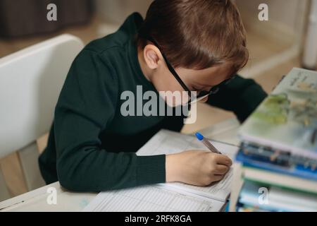 close up view of 7 eyars old caucasian boy wearing glasses doing homework at small desk behind pile of books Image with selective focus Stock Photo