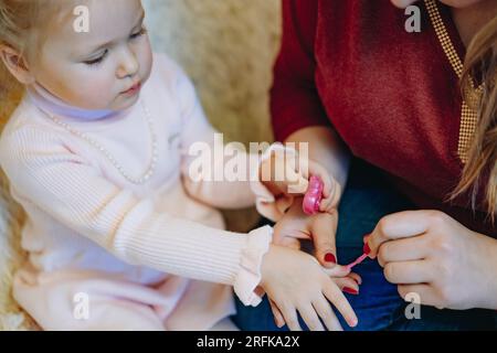 mother is coloring nails of her little girl with pink nail polish. Having fun making manicure at home Stock Photo