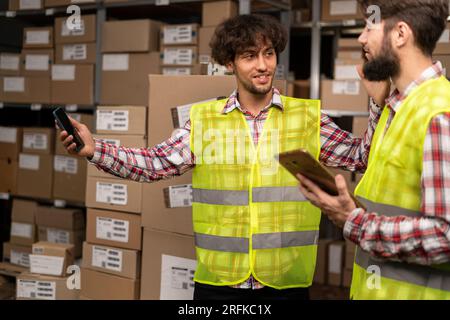 Two multiethnic warehouse workers discussing while standing near shelves with boxes Stock Photo