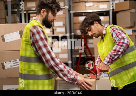 Two warehouse workers sealing cardboard boxes for shipping in a large warehouse. Stock Photo