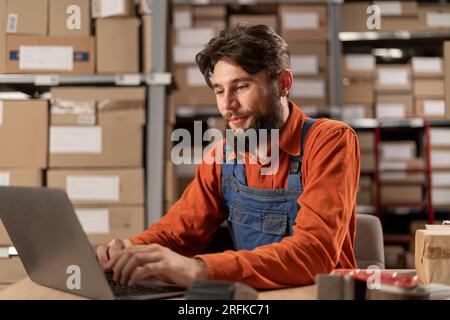Focused warehouse employee working on laptop in a large storehouse sitting at the table Stock Photo
