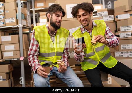 Two warehouse workers having lunch chatting and watching something on smartphone screen online Stock Photo
