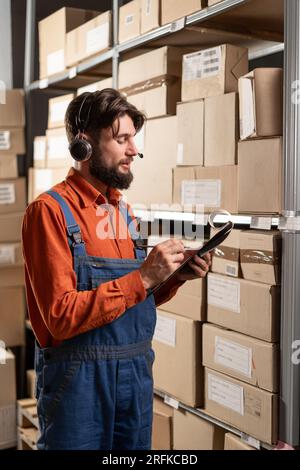 male worker wearing headset in logistics distribution warehouse writing notes in clipboard Stock Photo