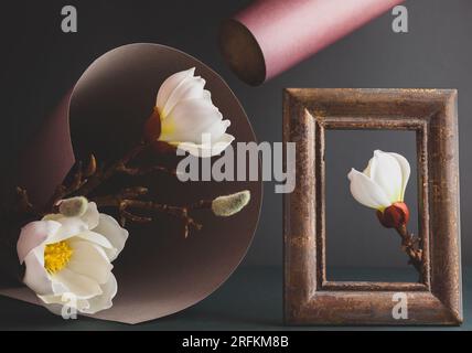 Creative still life with white magnolia, paper, vintage frame and dried branch of tree on dark background. Trendy nature concept. Stock Photo