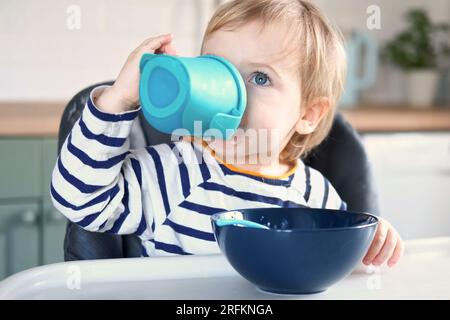 One year old hungry girl in striped casual clothes sits at white table in highchair, drinks water from baby bottle, sippy cup. Blurred dining room bac Stock Photo