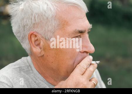 Senior man lighting up a cigarette outdoors against green nature  background. Old thoughtful man smoking, looking aside. Close Up Face.  Relaxed pension Stock Photo