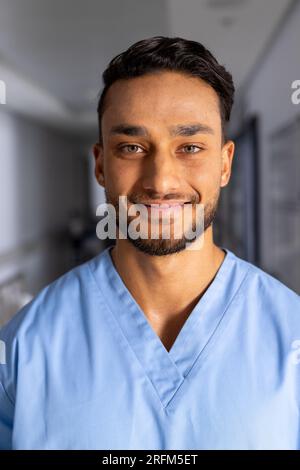 Portrait of happy biracial male doctor wearing scrubs in corridor at hospital Stock Photo