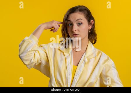 You are crazy, out of mind. Displeased woman pointing at camera, show stupid gesture finger near head, blaming some idiot for dumb insane plan idea, senseless talk. Girl isolated on yellow background Stock Photo