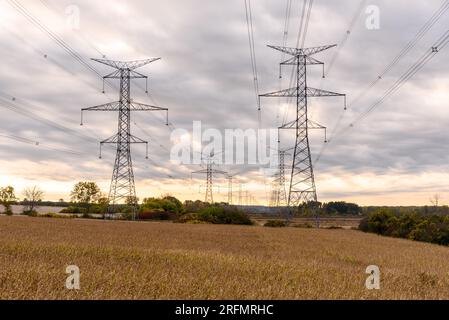 Electricity pylons supporting high voltage power lines in the countryside of Ontario under cloudy sky at sunset in autumn Stock Photo