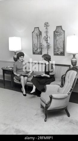 1950s, historical, two elegantly dressed young ladies sitting chatting together on a sofa, while browsing magazines, in a hotel lobby, USA. Stock Photo