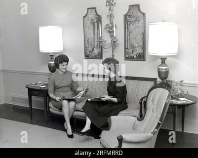 1950s, historical, two elegantly dressed young ladies sitting chatting together on a sofa, while browsing magazines, in a hotel lobby, USA. Stock Photo