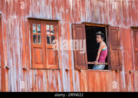 A young man with tatoos and dyed hair looks out of an open window of an orange wooden facade at Inle Lake in Myanmar Stock Photo