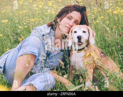Beagle dog portrait with its woman owner looking at camera while sitting in high green grass. Female hugging her pet friend and looks slightly sad. Ti Stock Photo