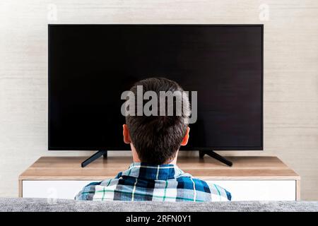 Back View Of Man Watching Television. Flat screen Plasma Set With Empty Screen. Sitting On Sofa At Home. Unrecognizable Guy Watching Movie On Weekend. Stock Photo