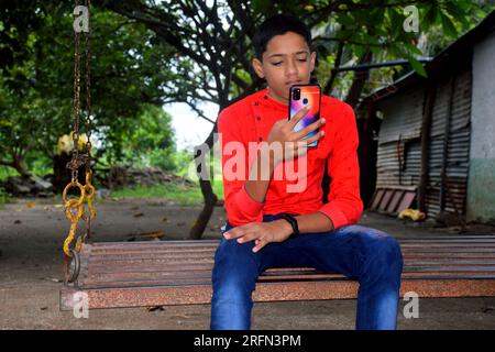 Indian boy in red shirt and blue jeans sitting in iron swing and using mobile. Garden background Stock Photo