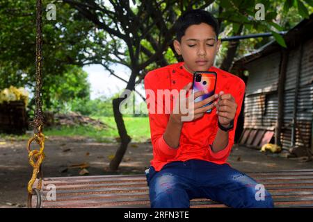 Indian boy in red shirt and blue jeans sitting in iron swing and using mobile. Garden background Stock Photo