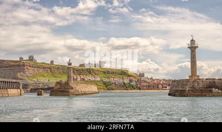 A panorama of Whitby from the sea. A pair of lighthouses guard the entrance to a harbour. The Abbey and a church are on the cliff and people are on a Stock Photo