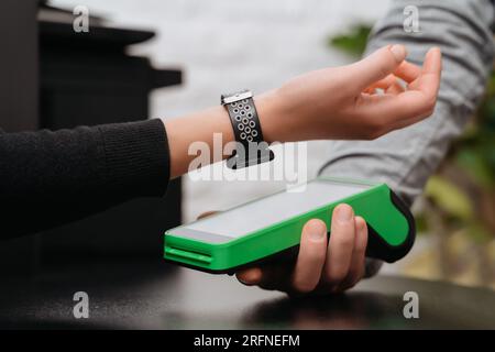 Client making contactless payment by smart watch with NFC technology. Close-up of modern woman with hand watch making express payment via nfc terminal while buying clothes in store. Stock Photo