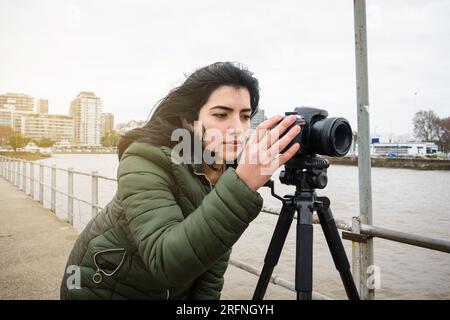 young latin woman in green jacket, content creator, is standing on the pier on a cloudy day, checking the video she made with her digital camera, tech Stock Photo