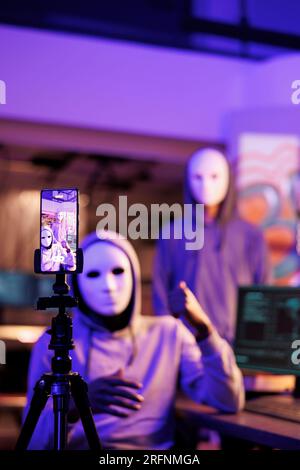 Smartphone screen with hackers recording threat video, asking victim for sensitive information and money. Criminals wearing anonymous masks threatening online and streaming fraud at night time Stock Photo