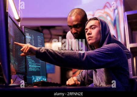 Hacker pointing at malware on screen while working together in abandoned warehouse. Asian criminal in hood showing code lines on computer to ransomware developer while cracking password Stock Photo